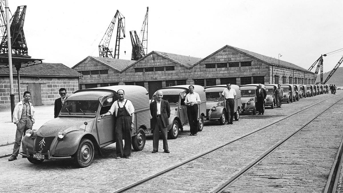Trabajadores junto a automóviles de Citroën Hispania y los almacenes de Zona Franca en el puerto, en 1958