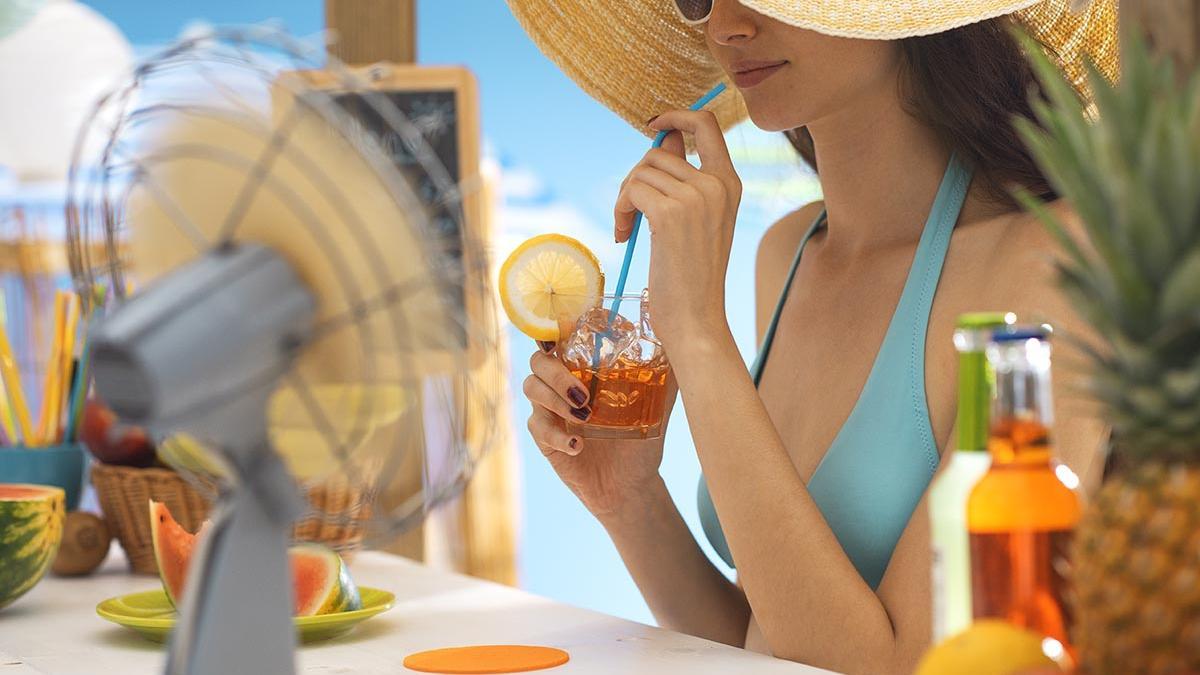 Ventiladores para combatir el calor, una joven en bikini con sombrero bebe un refresco en la playa