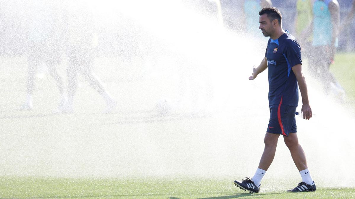 Xavi observa el entrenamiento mientras los aspersores mojan el césped.