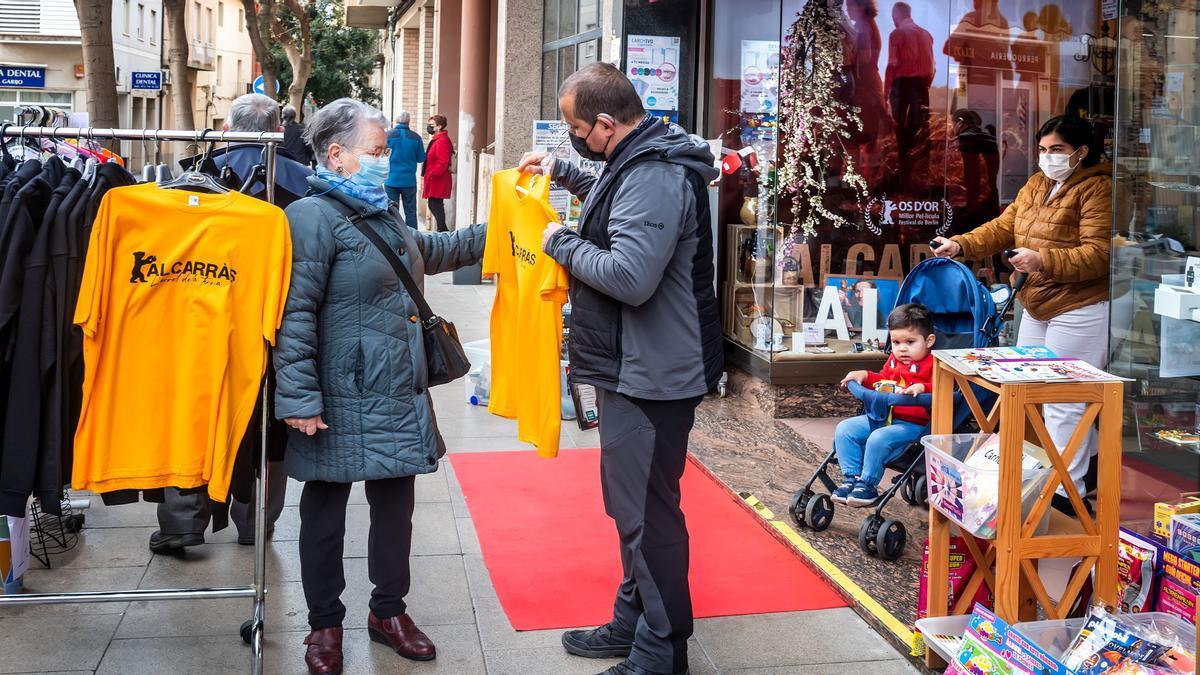 En día de mercado en la calle, Avel·lí Estela enseña a Fina, vecina del municipio, las camisetas que se han confeccionado con el nombre de Alcarràs y un oso de oro que sostiene una manzana. El quiosco Novetats Moliner, de la calle mayor de Alcarràs, ha decorado su escaparate con merchandising de Alcarràs a propósito del premio del Oso de Oro a la película del mismo nombre. En la puerta del comercio han puesto una alfombra roja cinematográfica, por iniciativa de la asociación de comerciantes. Alcarràs, Segrià. 4 Marzo 2022 Foto Jordi V. Pou