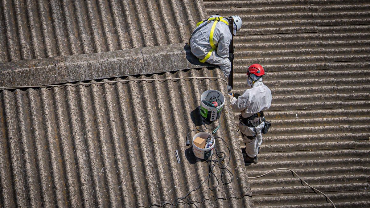 Dos trabajadores, con máscaras y trajes protectores, sobre el tejado con fibra de amianto.