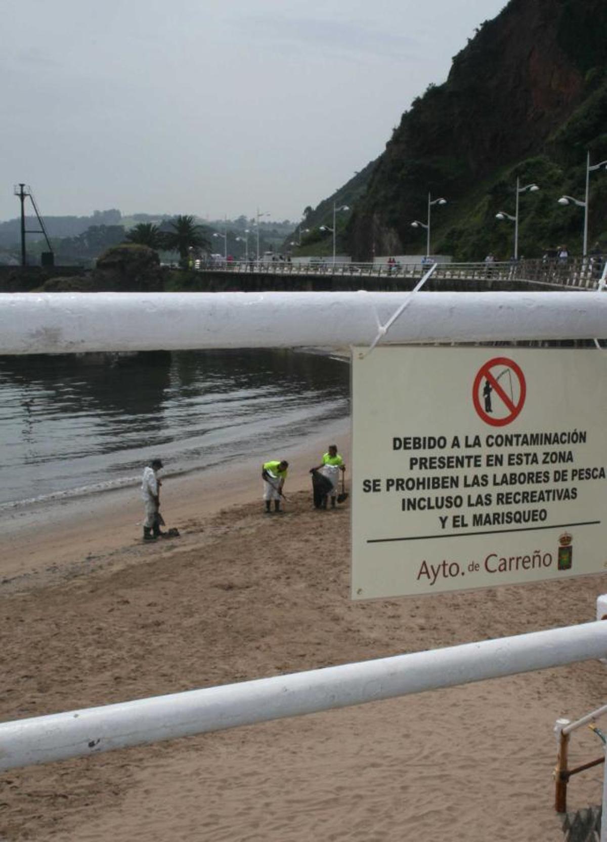 Un cartel advierte de las restricciones pesqueras mientras los operarios limpian la playa de Candás en los días posteriores al vertido.