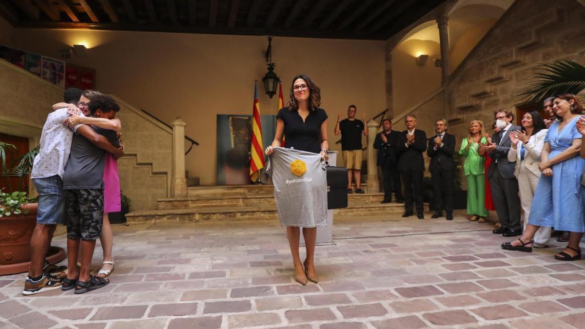 La vicepresidenta de la Generalitat, Aitana Mas, muestra la camiseta que le regaló Mónica Oltra, a la izquierda de la imagen abrazando a sus hijos, con el aplauso del Consell.