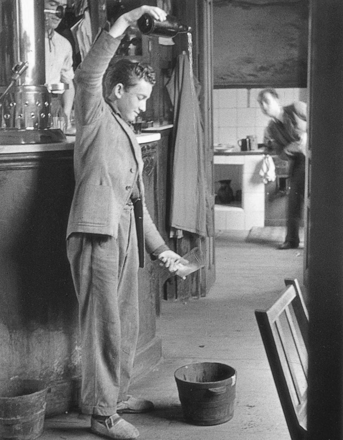 Un hombre escancia sidra en el café de Colasín en 1948, con el brazo algo doblado y el vaso colocado en un lateral. | Muséu del Pueblu d’Asturies /  Colección Valentín Vega