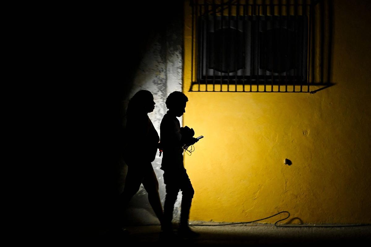 A woman holds a flashlight while walking with a man on a street during a blackout in Havana on March 16, 2026. Cuba suffered a widespread power cut on March 16, 2026, according to the national electricity company, against the backdrop of a severe crisis on the island caused by the US energy blockade. (Photo by Yamil LAGE / AFP)