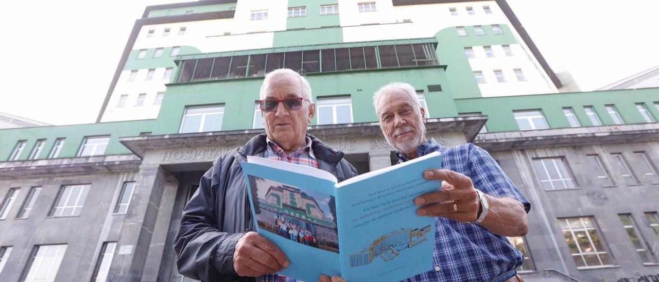 José Miguel Estevan Solano, a la izquierda, y José Enrique Menéndez Menéndez, delante del Hospital General de Asturias, con un ejemplar del libro elaborado para la reunión de médicos residentes. | Miki López