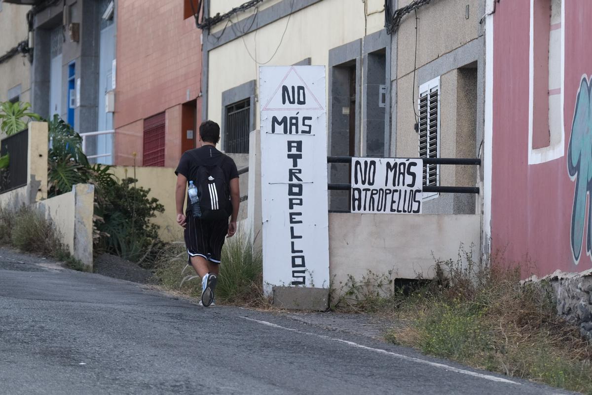 Un joven camina por la carretera de Almatriche junto a un cartel en el que se reclama la construcción de las aceras.
