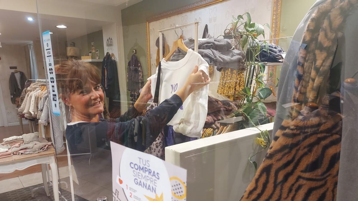 Ana Suárez coloca ropa en su tienda tras un escaparate con el cartel del programa "Gijón compra y vuelve"