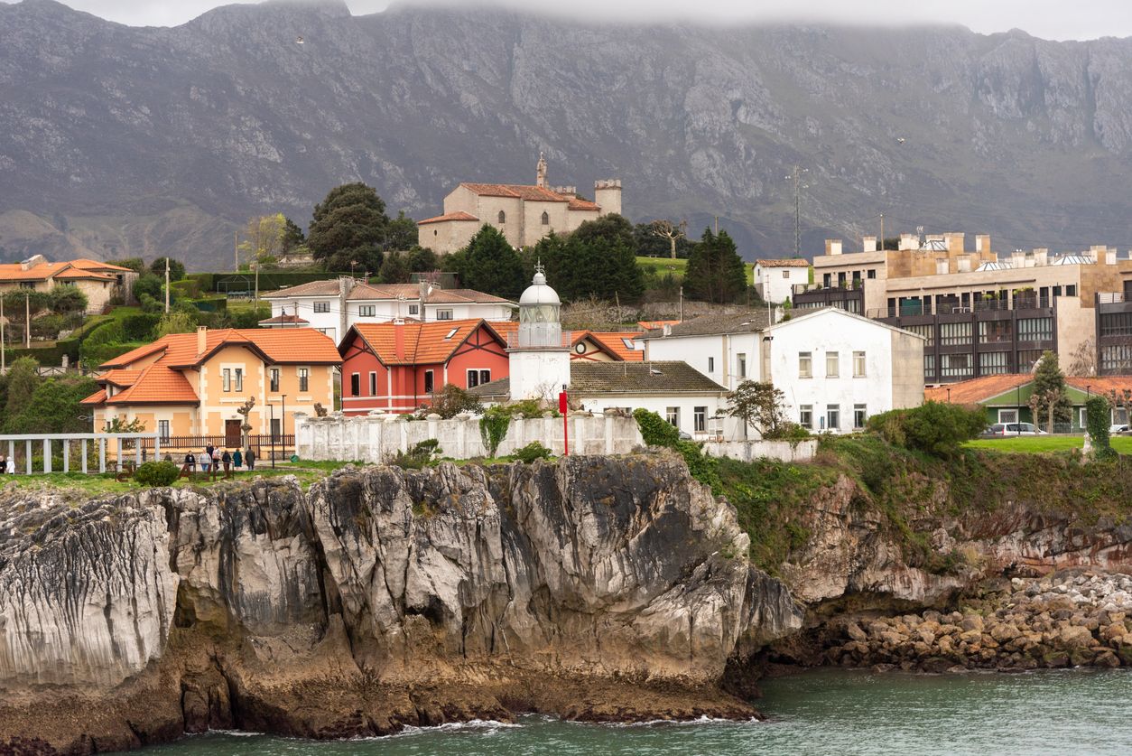 Vista panorámica del faro de la villa turística de Llanes junto al mar y sobre los acantilados rodeados de pequeñas casas y montañas rocosas al fondo durante un día nublado en Asturias
