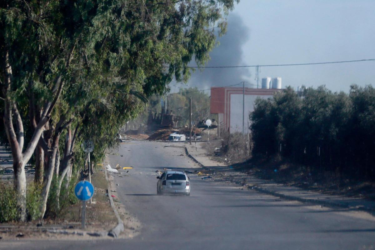 Un coche conduce por una calle solitaria en la ciudad de Gaza. Al fondo, columnas de humo.