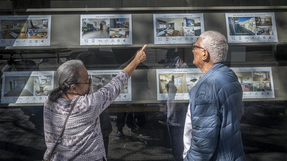 Dos personas miran carteles de pisos de alquiler y compra en una inmobiliaria de Barcelona.