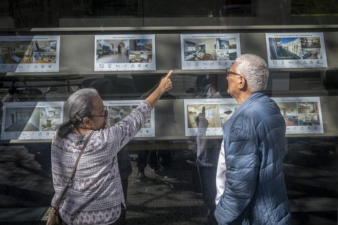 Dos personas miran carteles de pisos de alquiler y compra en una inmobiliaria de Barcelona.