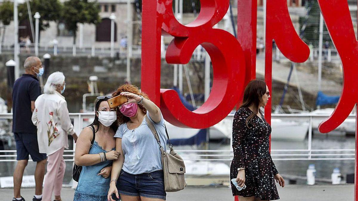 Unas turistas con mascarilla se hacen una foto delante de las &quot;letronas&quot; de Gijón.