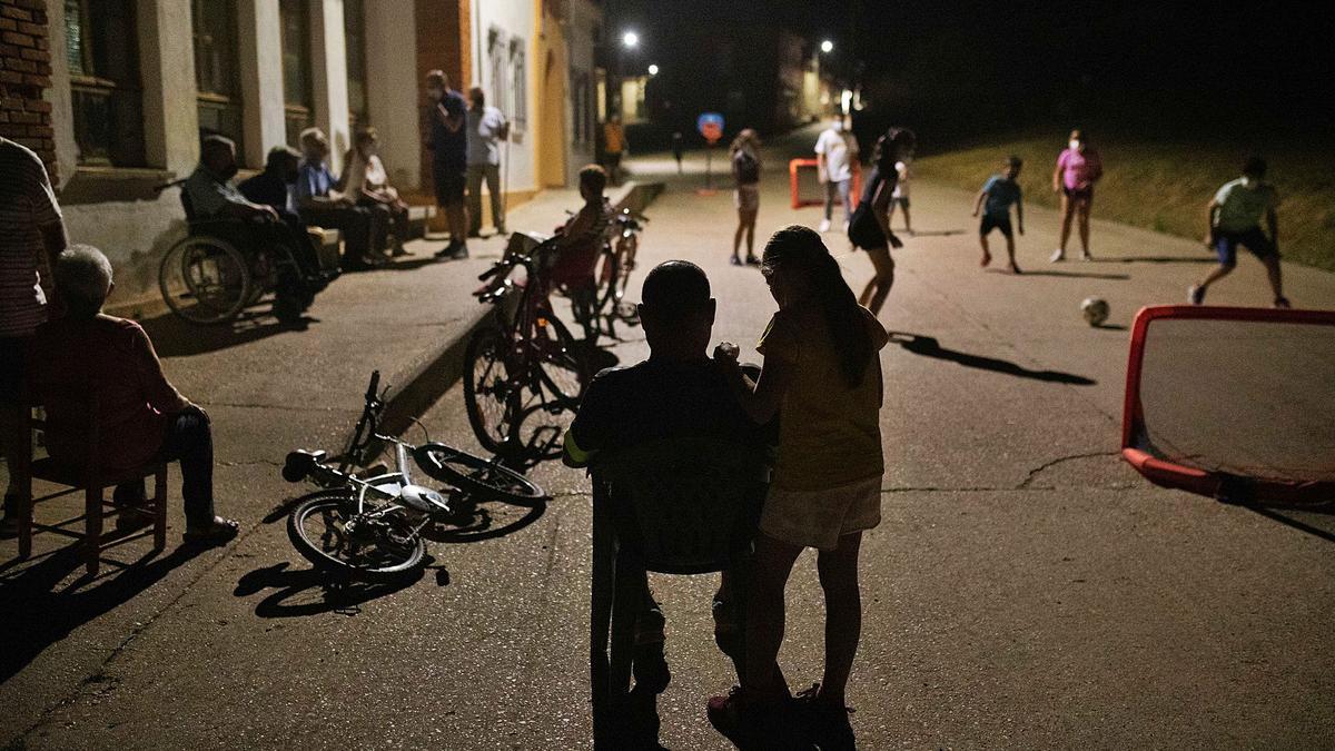 Un grupo de personas toma el fresco ante las escuelas de Videmala mientras observa un partido de fútbol infantil.