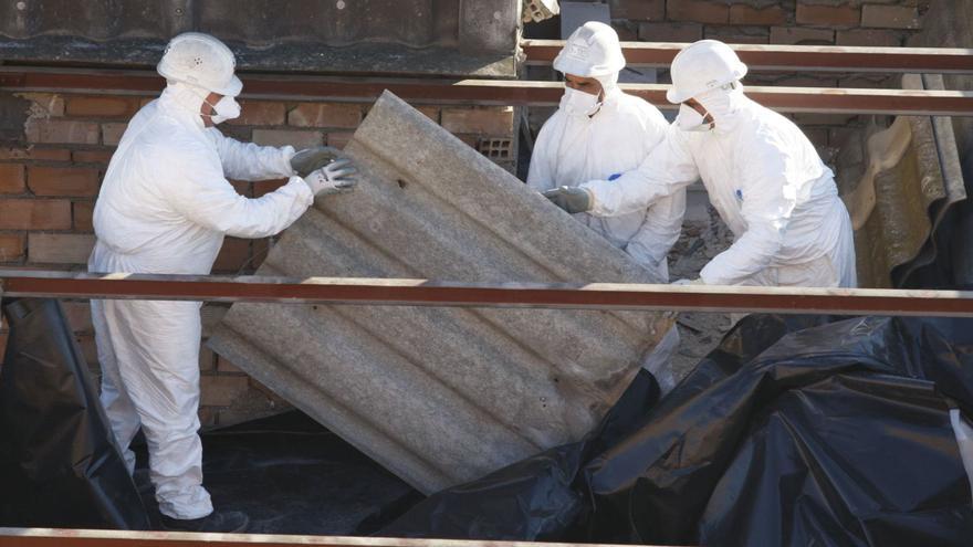 Tres operarios retiran amianto del tejado de un edificio protegidos con los trajes y máscaras de seguridad debido a su toxicidad. | L.O.