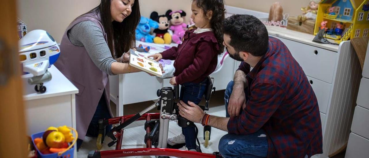 Alejandra, con el soporte que necesita para mantenerse en pie, mira un cuento junto a sus padres.