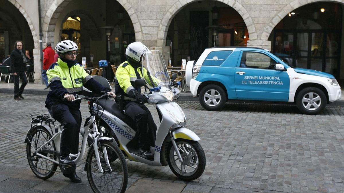 Una agent cívic i un policia passen per davant d’un vehicle del servei de teleassistència. | MARC MARTÍ