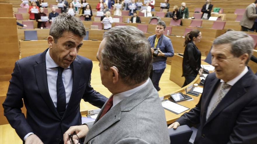 El presidente Mazón y el vicepresidente Gan conversan en el pleno con los diputados de la oposición al fondo mostrando carteles con la cifra de fallecidos. |