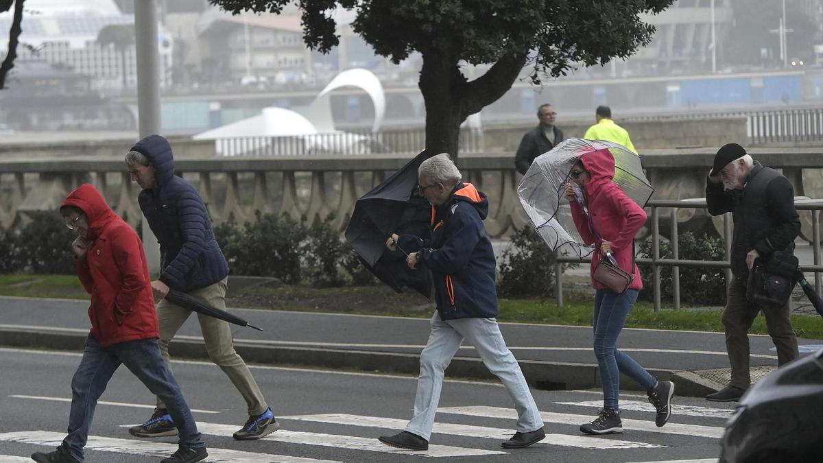 Peatones cruzan con viento y lluvia en el paso frente a la coraza de Riazor.