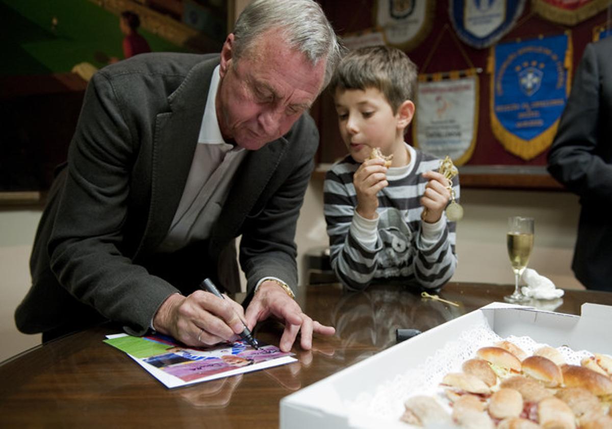 El seleccionador català Johan Cruyff firma un autògraf supervisat per un nen mentre menja uns canapès en la presentació de la selecció catalana que jugarà el dia 30 contra Tunísia .