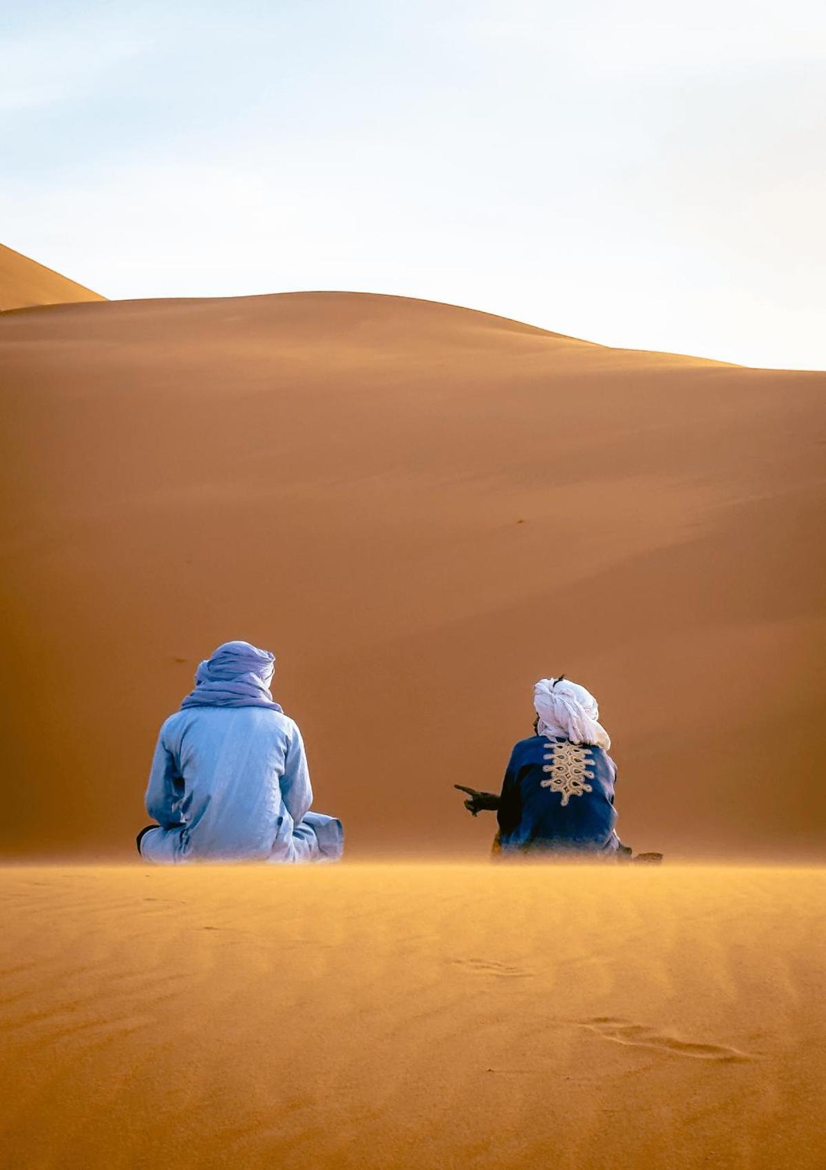 Erg Chebbi, Marruecos. Dos guías bereberes conversan sentados sobre una duna del Sáhara. Las dunas de Erg Chebbi, entre las más altas de Marruecos, alcanzan los 150 metros y son conocidas también como "las dunas de Merzouga". (iPhone 16 Pro Max)