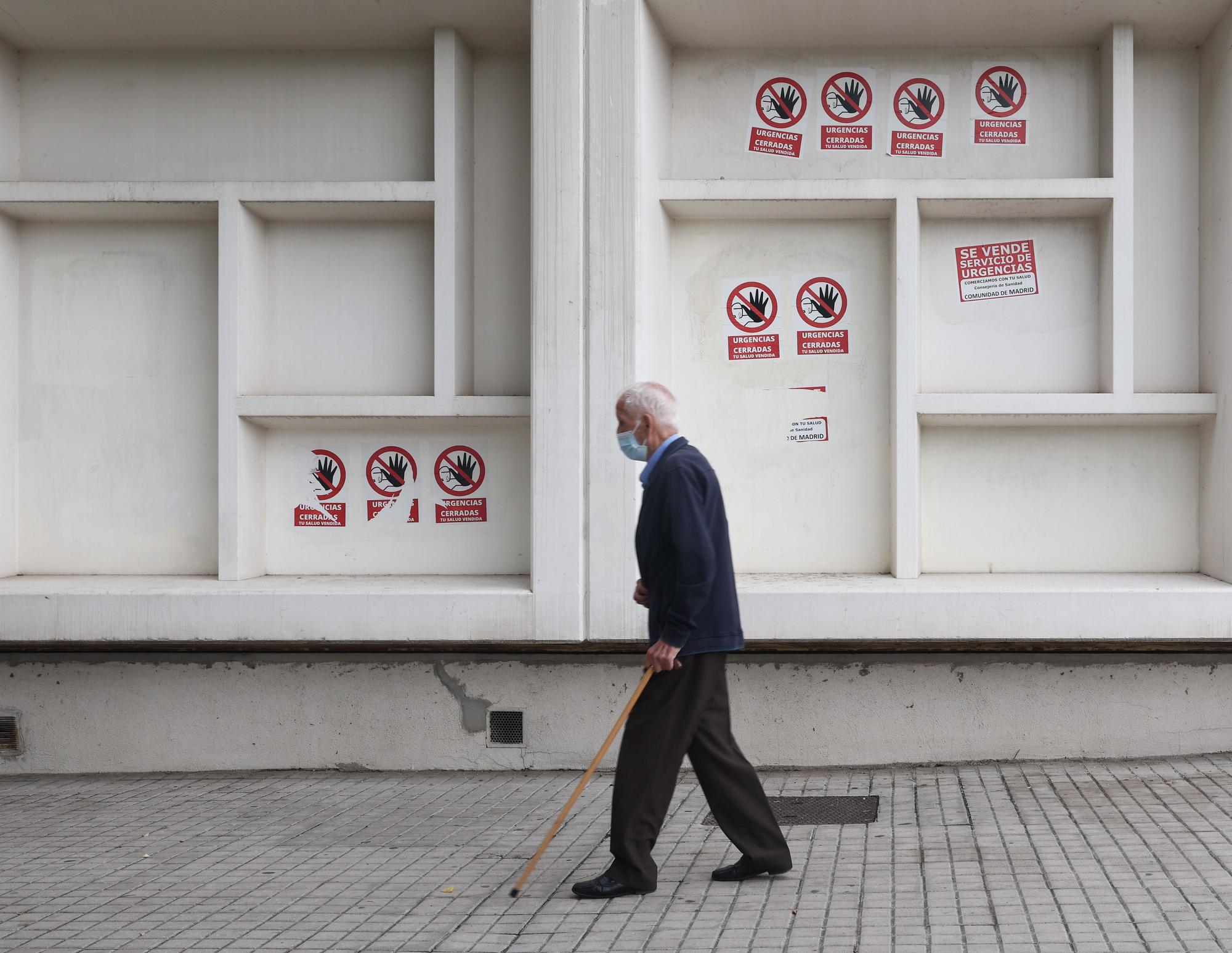 Un hombre mayor con mascarilla y bastón camina delante del Centro de Salud Pavones, donde hay carteles de protesta que rezan 'Urgencias Cerradas, tu salud vendida', a 21 de octubre de 2022, en Madrid (España).