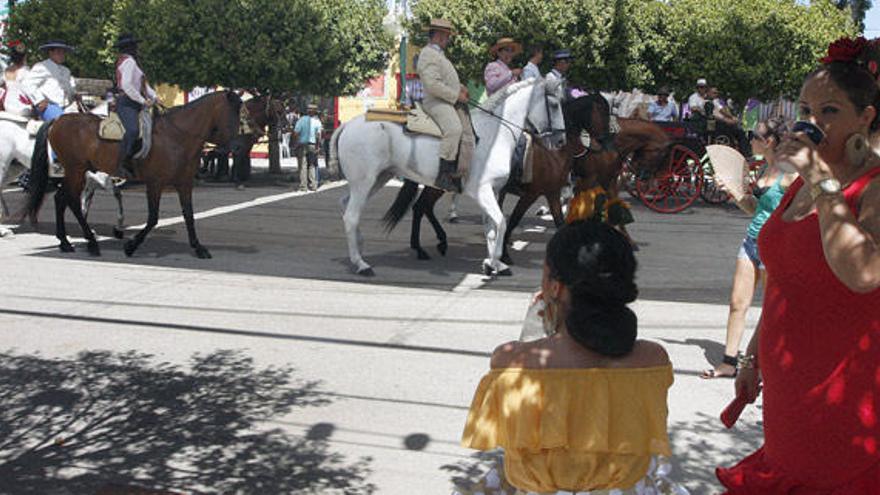 Dos mujeres, ataviadas con el traje de flamenca, intentan refrescarse a la sombra del calor de agosto mientras observan pasear a los caballistas.