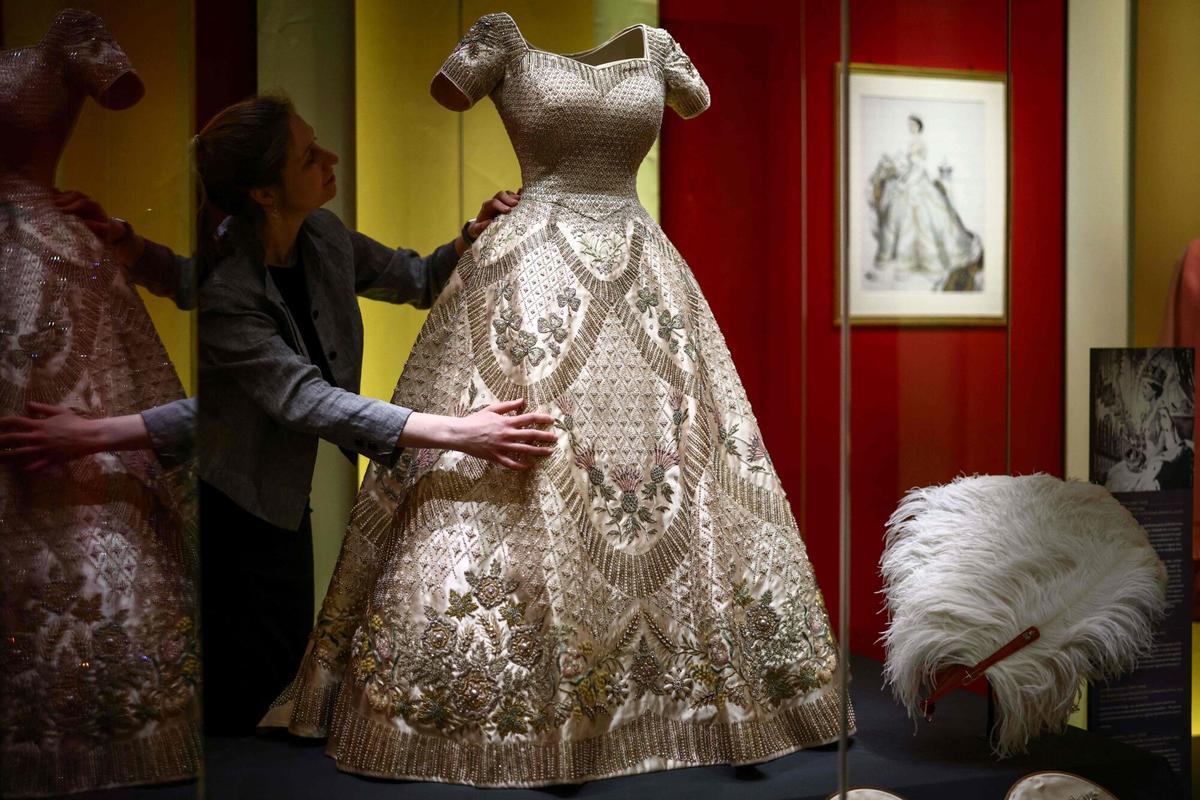 A member of the Royal Collection Trust staff poses with a 'The Coronation Dress' (C) from 1953, during a media preview of the exhibition 'Queen Elizabeth II: Her Life in Style' at the King’s Gallery in Buckingham Palace in London on April 9, 2026. The largest exhibition showcasing Queen Elizabeth II’s fashion ever staged is being held from April 10 to October 18, 2026. (Photo by Henry NICHOLLS / AFP) / RESTRICTED TO EDITORIAL USE - NO MARKETING - NO ADVERTISING CAMPAIGNS. RESTRICTED TO EDITORIAL USE - NO MARKETING - NO ADVERTISING CAMPAIGNS