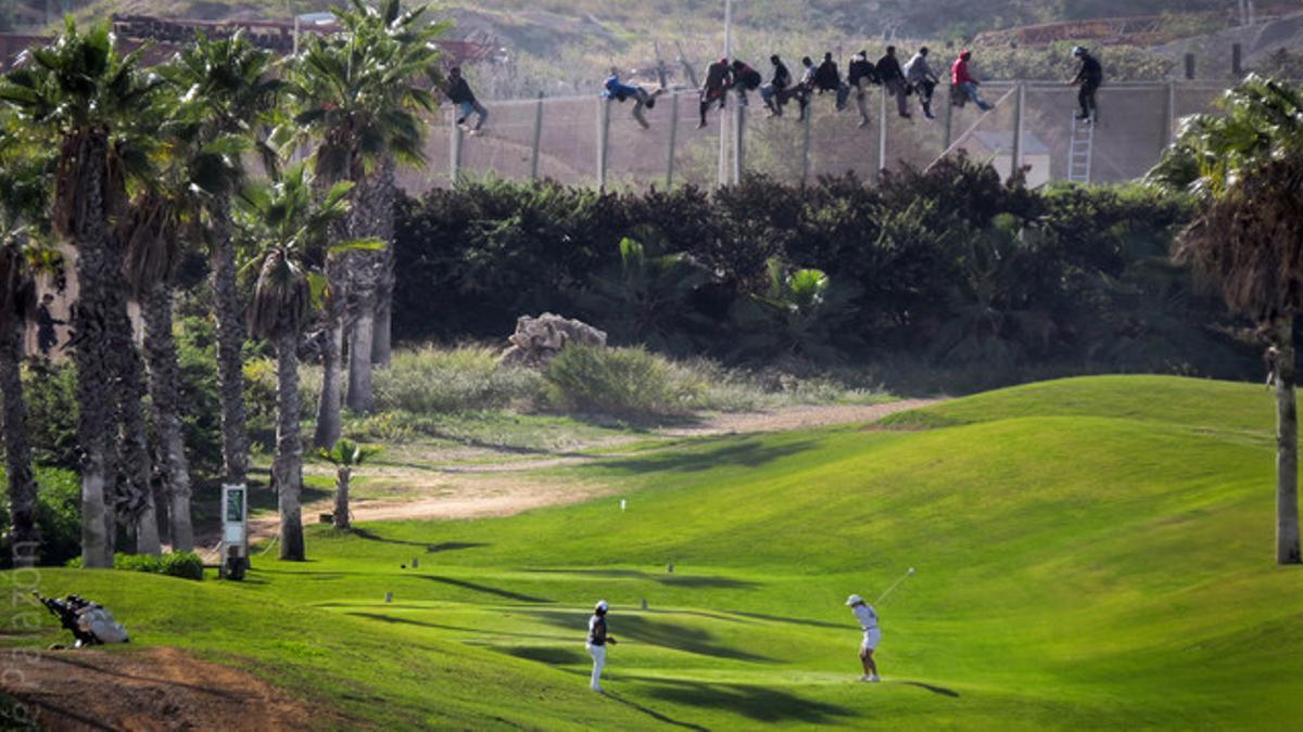 Un grupo de inmigrantes tratan de saltar la valla de Melilla junto a un campo donde juegan dos golfistas.