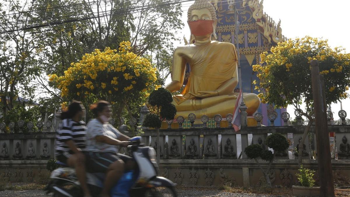 Dos tailandeses circulan en moto delante de una estatua de Buda con mascarilla en las afueras de Bangkok.