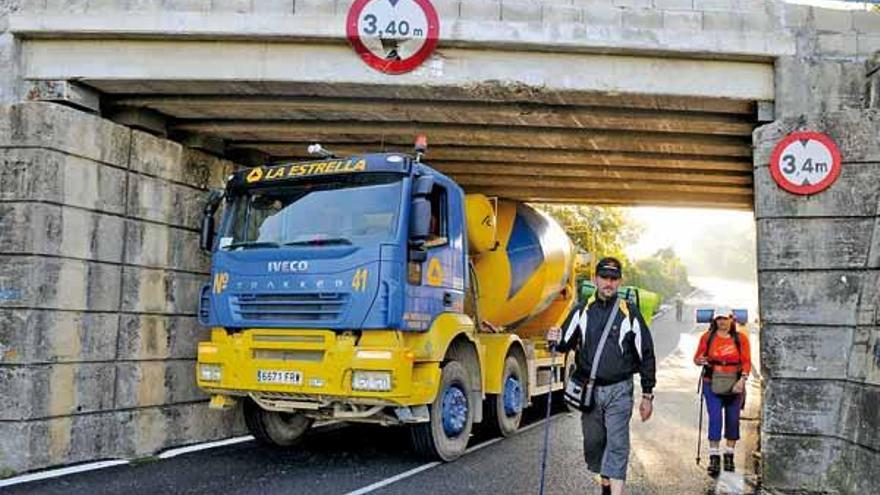 La hormigonera encajonada bajo el puente con la señal de gálibo encima mientras unos peregrinos pasan a su lado.