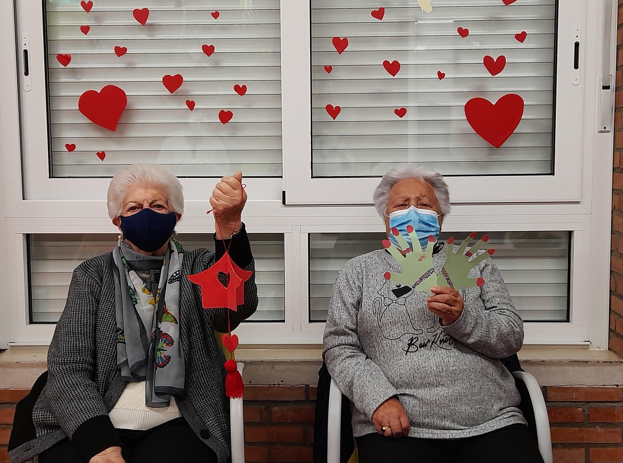 Dos usuarias muestran sus trabajos ante una ventana repleta de corazones, elaborados por San Valentín.