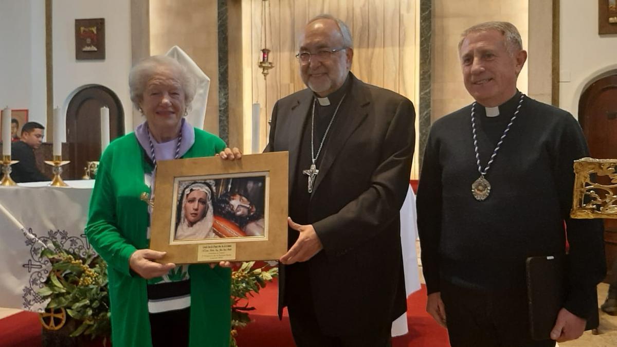 La hermana mayor de la Cofradía, Rosario Menéndez, entrega una placa con las imágenes del Cristo Yacente y la Señora de la Soledad al Arzobispo, en presencia del párroco de Lugones, Joaquín Manuel Serrano.