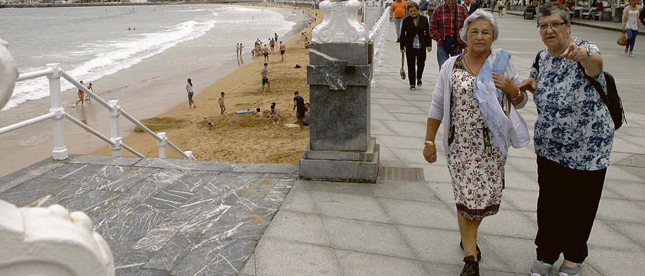 Dos mujeres pasean por el muro de San Lorenzo, en Gijón, con chaqueta, mientras la playa permanece semivacía.