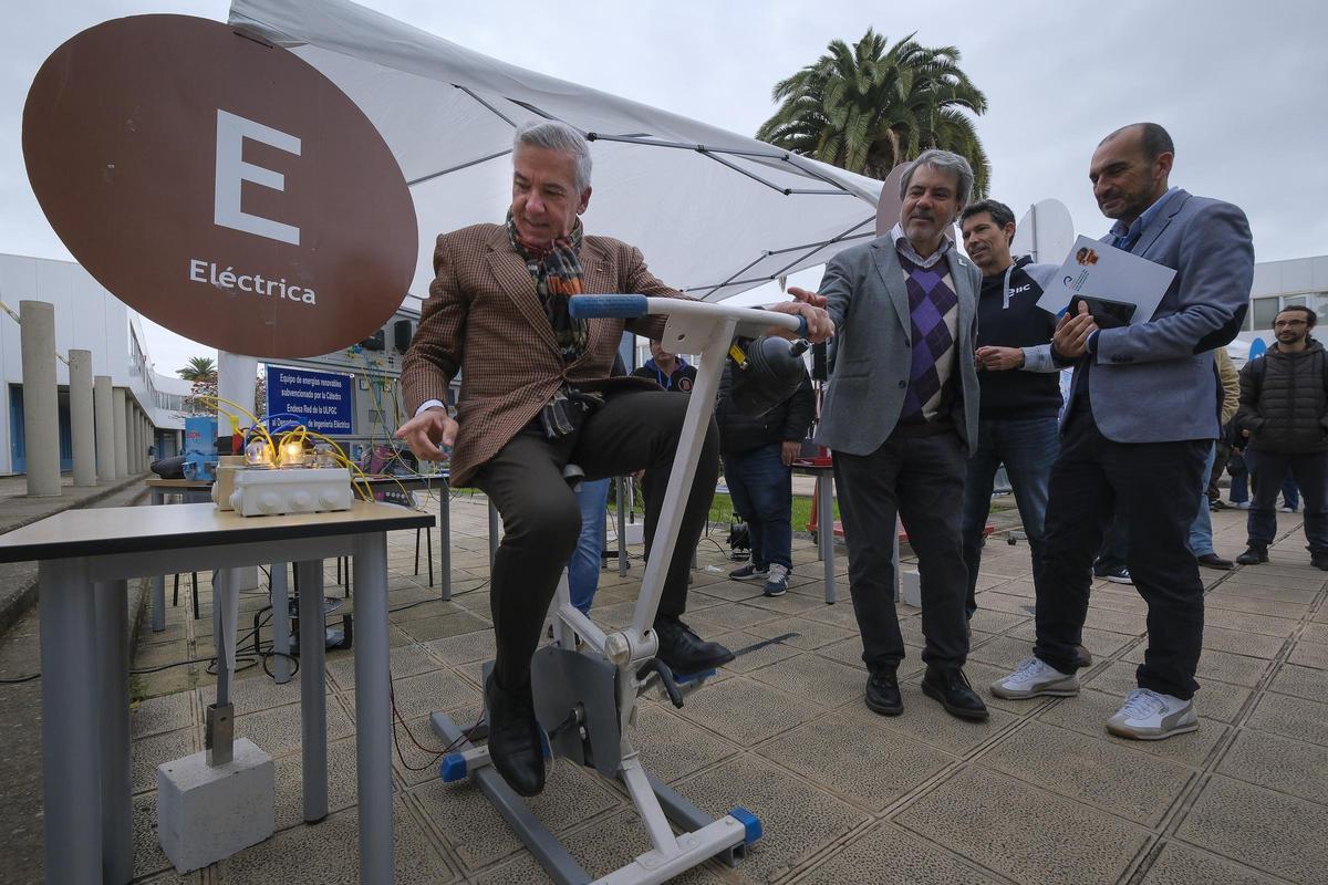 El rector de la ULPGC, Lluís Serra, pedalea en una bicicleta para encender una bombilla mediante la cinética en el stand de Ingeniería Eléctrica.