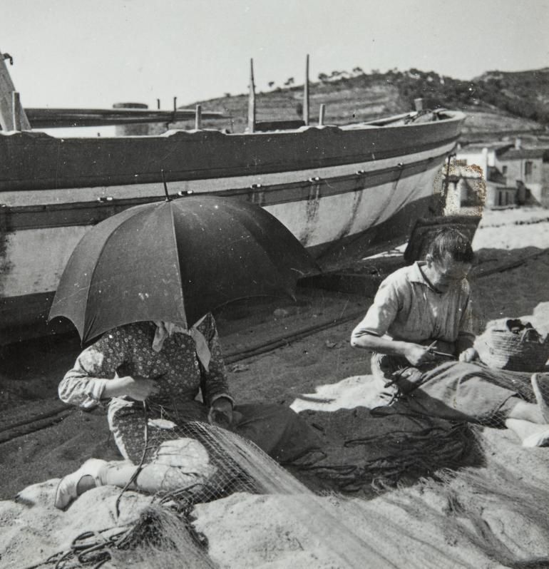 Dones cosint xarxes de pesca a la platja, al costat de les barques.