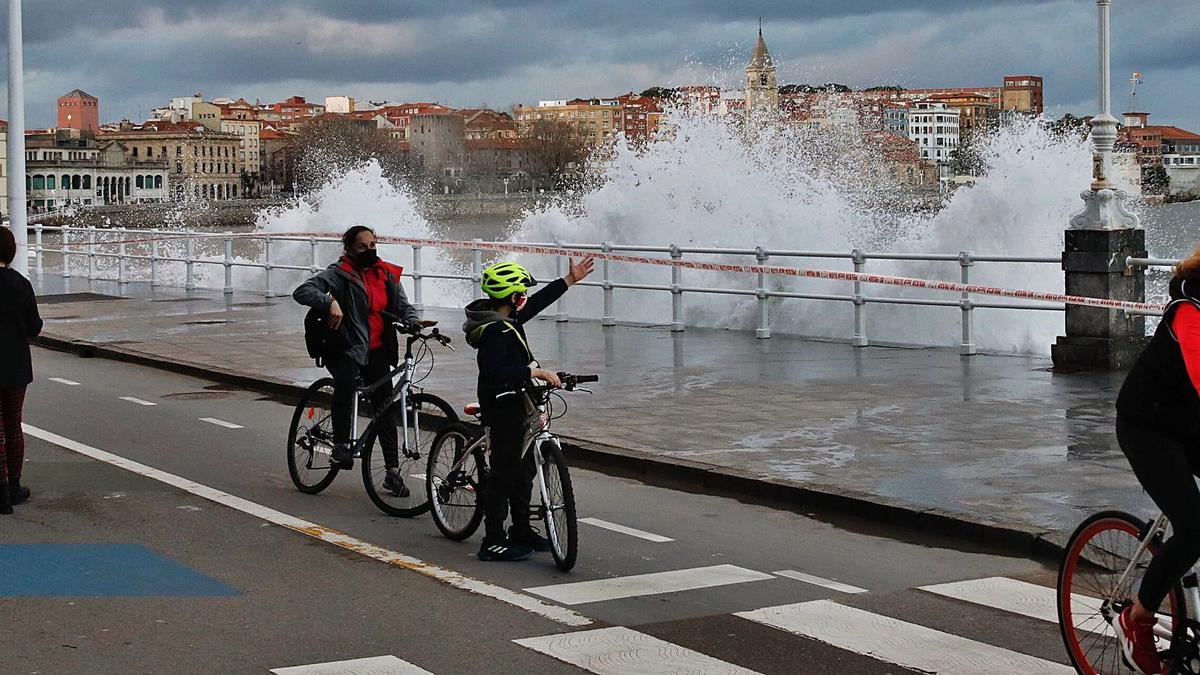 Una ola sacude el paseo del Muro ante varios ciclistas y paseantes.