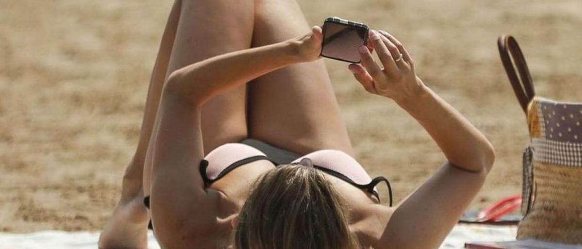 Una mujer mira su teléfono móvil mientras toma el sol en una playa.