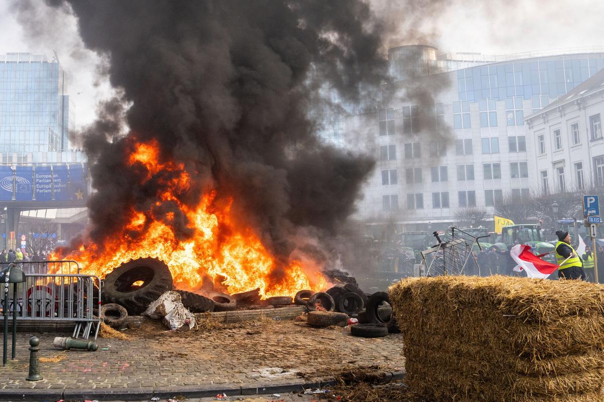 Un manifestante ondea una bandera entre neumáticos en llamas frente al Parlamento Europeo. La movilización, convocada por agricultores europeos, denuncia los recortes en los subsidios de la PAC y el acuerdo comercial con Mercosur por temor a la competencia desleal. 18 de diciembre de 2025, Bruselas.