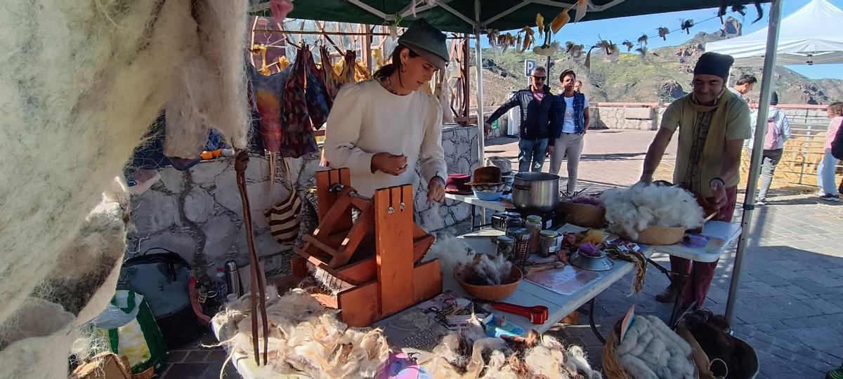 La artesana Lesley Bohncke, preparando la lana para confeccionar alguna prenda, con algunos utensilios para su trabajo, un caldero al fuego para teñir la ropa, y los chalecos ya acabados expuestos a su espalda. A su lado, Pablo, limpiando la lana.