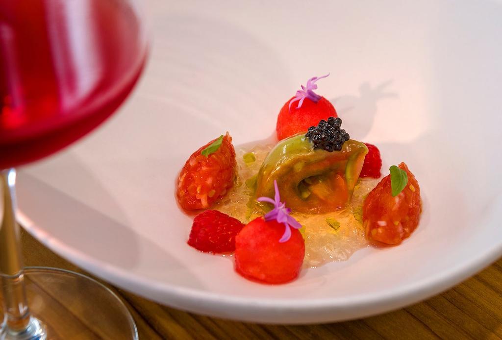Tomate de verano: sobre una cama de agua de tomate gelificado colocar el tomate pelado con caviar, dos quenelles de tartar de tomate, frambuesas y bolitas de sandía; aliñar con aceite, sal y vinagre. Decorar con flores de ajo y hojas de albahaca.