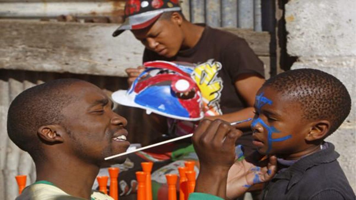 Un artista pinta la cara d’un nen mentre un company realitza unmakarapa --una mena de casc retallat i pintat a mà que és tradicional en el futbol sud-africà--, a la població de Port Elisabeth.