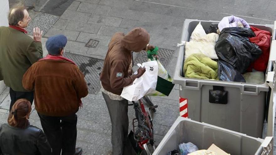 Varios transeúntes pasan junto a un hombre que rebusca en los contenedores de basura.