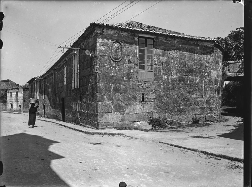 Vista lateral de construcción típica en piedra con miradores cerrados y escudo en la fachada lateral. Tendido eléctrico. En la calle, mujer de negro con cesto de ropa en la cabeza] otto.jpg