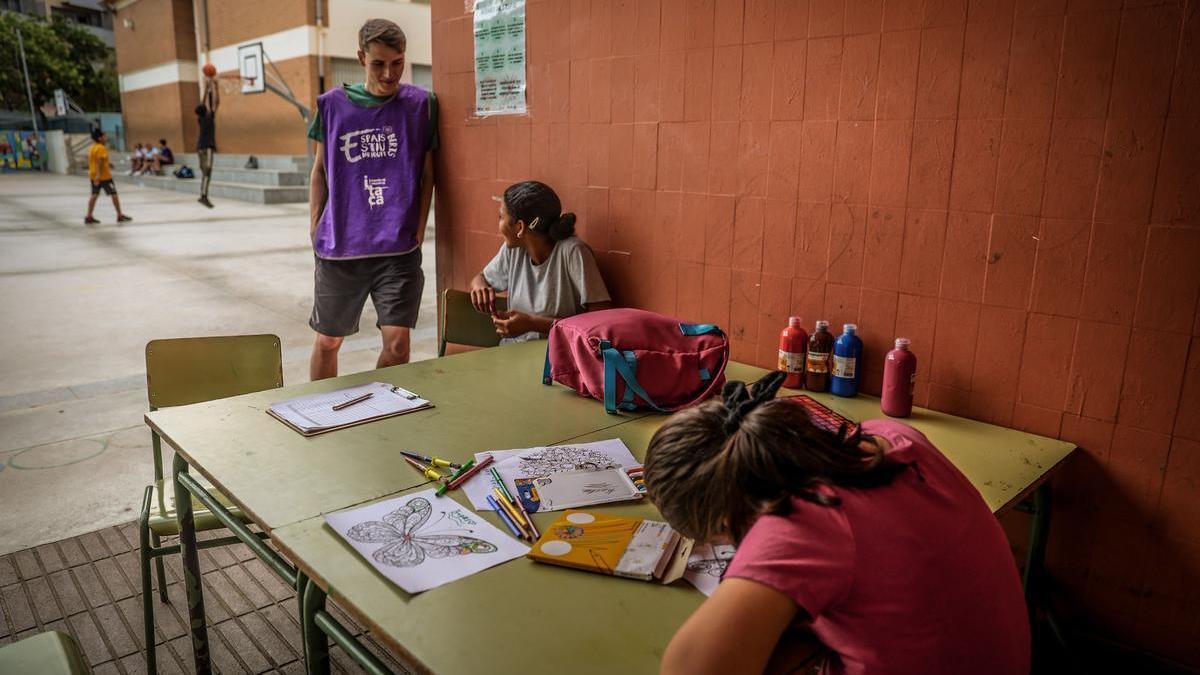 Dos niñas realizan manualidades, ante la mirada de un educador, en la escuela Pau Casals de l’Hospitalet, cuyo patio permanece abierto todo el verano patra actividades de ocio gratuitas.