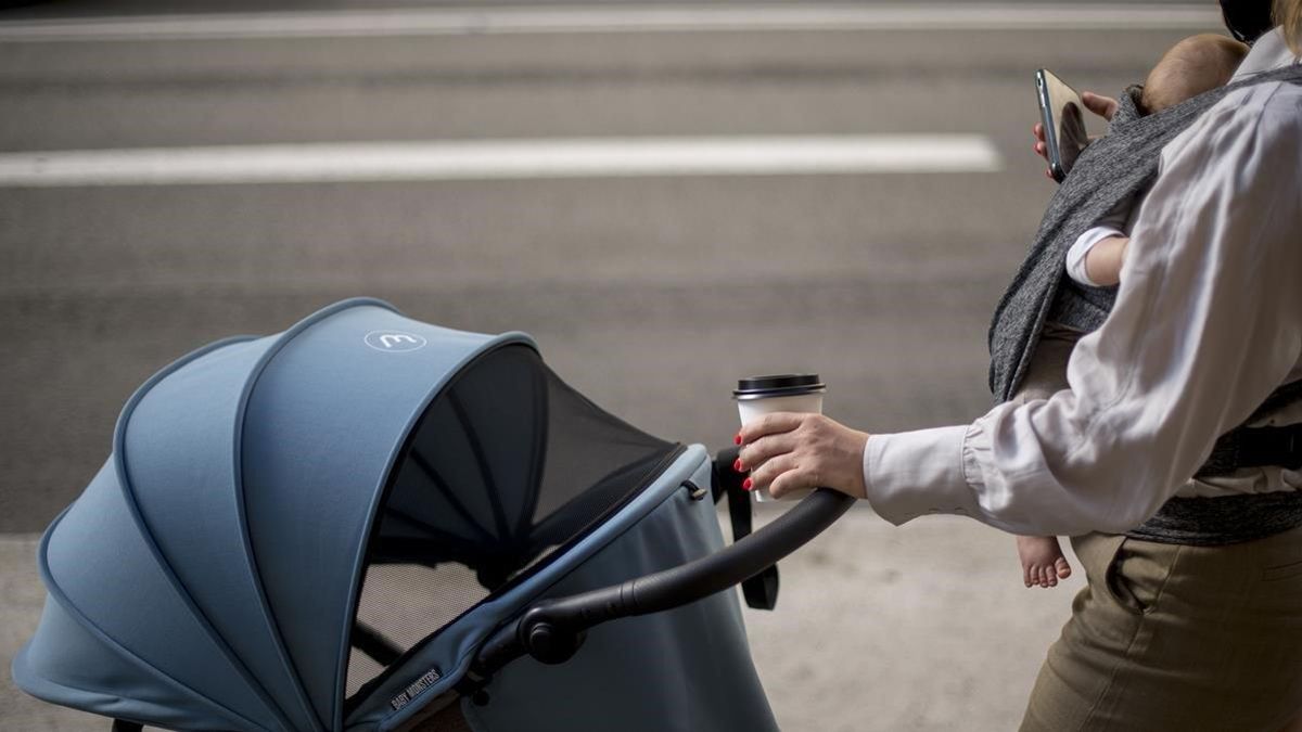 Una mujer con mascarilla consulta el móvil con su bebé a cuestas mientras toma un café para llevar y empuja un carrito, el 31 de agosto del 2020