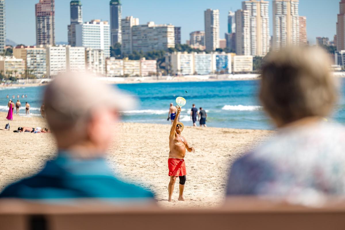 Un jubilado juega a las palas en la playa de Benidorm, mientras otros le observan desde el paseo.