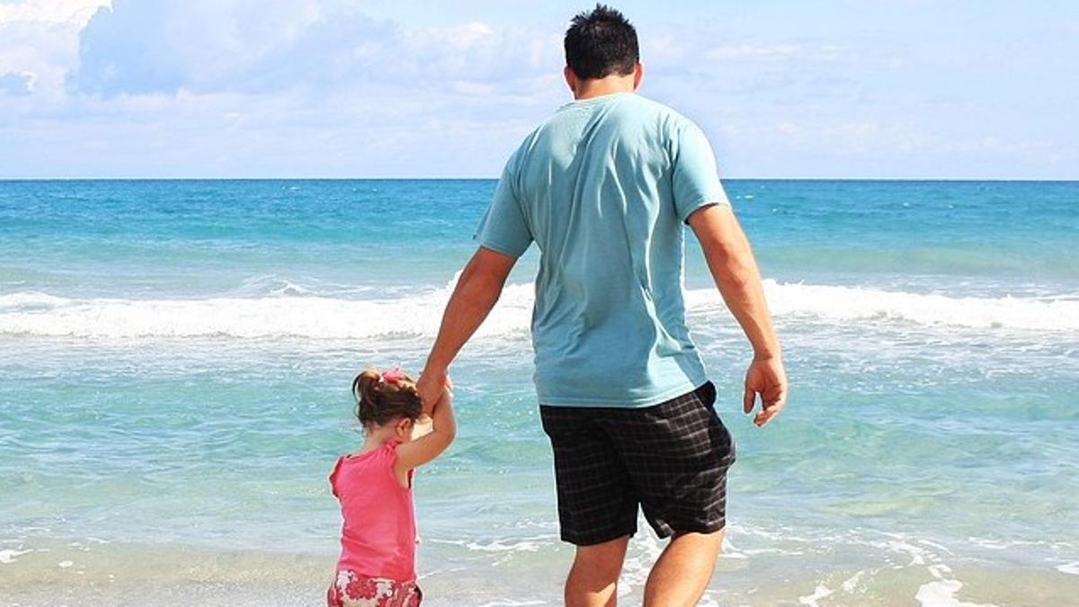 Un padre lleva de la mano a su hija por una playa.