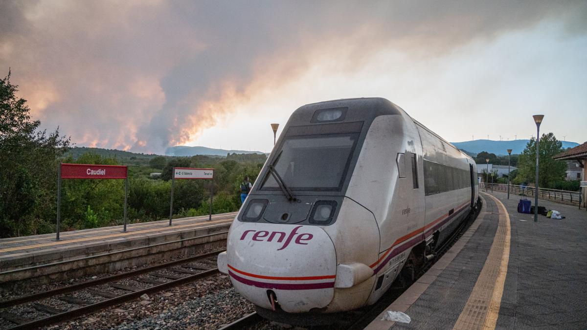 El tren afectado, en la estación de Caudiel tras ser evacuado, con las llamas del incendio al fondo.