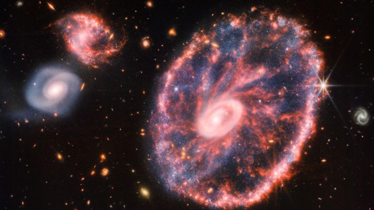 Una gran galaxia rosada que se asemeja a una rueda con un pequeño óvalo interior. A la izquierda, dos galaxias espirales más pequeñas del mismo tamaño.